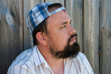 A young man with a beard in the hat on a wooden background wall looks thoughtfully into the distance