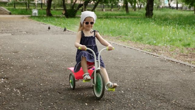 A little girl in sunglasses and a blue dress rides a pink tricycle on a track in a city park on a sunny summer day slow motion.