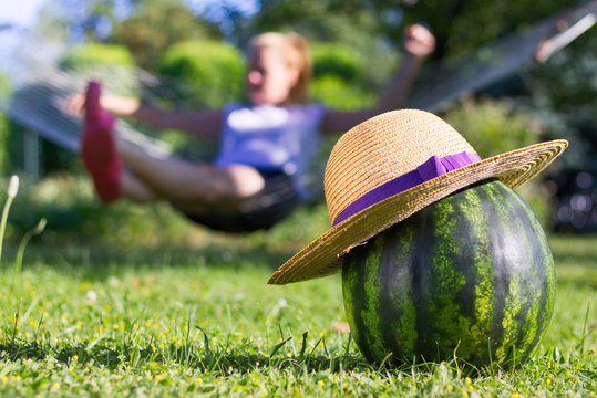 Happy summertime in the garden. A straw hat on a watermelon and a girl relaxing in a hammock in the background. 