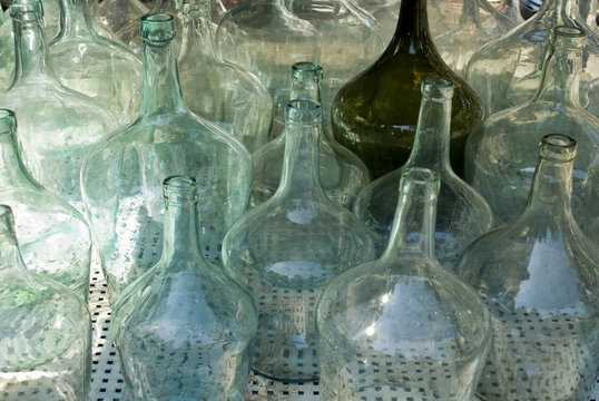 Closeup of a group of old damigiane (demijohns, carboy) glass for wine, of different colors, transparent white, green, brown, in a market in italy