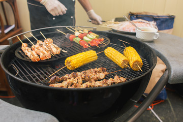 Corn, meat, vegetables on skewers cooked at barbecue outdoors with chefs hands out of focus. Street food, fast food