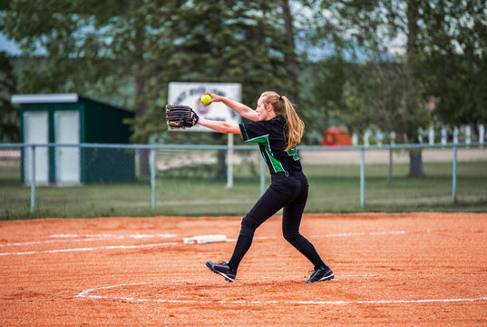 A teenage girl in fast ball pitching stance on the pitchers mound in black and green uniform