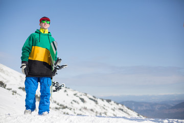 Snowboarder wearing snowboarding hoody sports gear posing with his snowboard looking away enjoying a sunny day at the mountains. Activities leisure lifestyle, winter sports concept
