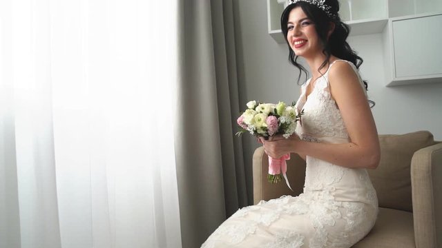 charming young bride is sitting on armchair in a wedding dress with a big bouquet of flowers, daydreaming