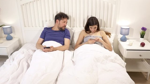 Jealous husband watching in bed wife writing text message with cell phone wide shot
