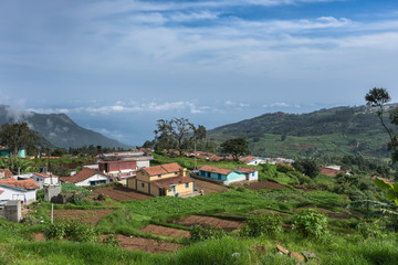 Fototapeta na wymiar Nilgiri Hills, India - October 26, 2013: Thalaikunha hamlet with small farms and green and brown vegetable plots under blue sky. Highland scenery with mountains, dark green forests, colorful houses