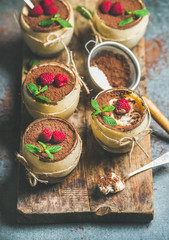 Homemade Italian dessert Tiramisu in individual glasses with mint leaves, fresh ripe raspberries and cocoa powder on rustic wooden board over grey concrete background, selective focus