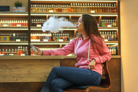 Vape woman. Portrait of young cute girl in pink hoodie smoking an electronic cigarette in the bar.