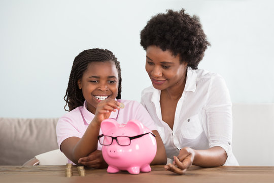 Girl With Mother Inserting Coins In Piggy Bank