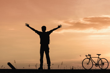 Silhouette of man raised his hands with the bike on the road at sunset