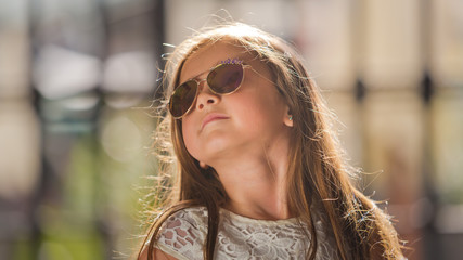 Portrait of a little girl in a white dress in sunglasses near the trellis fence. Close-up.
