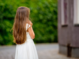 Naklejka premium Cute girl in white dress is standing with her back on a green fence background.
