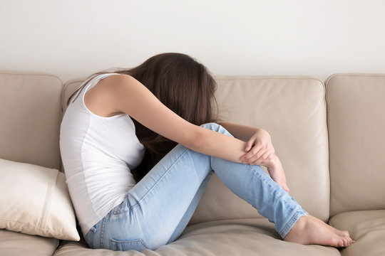 Depressed young woman sitting on sofa embracing knees with hands. Stressed and upset adolescent girl feels emotional emptiness, loneliness, worried because of relations. Teens psychological problems