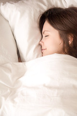 Young attractive woman sleeping in soft bed under warm blanket. Teenage girl falling asleep in bedroom after night party, seeing pleasant dreams in soft bed with fresh white sheets. View from above