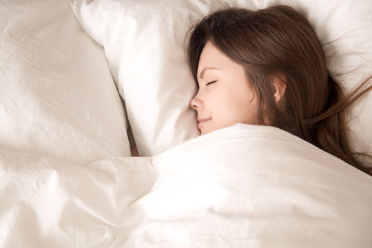 Young woman lying under warm blanket, seeing sweet dreams and smiling. Lady sleep well in bedroom with white sheets, relaxing in comfortable soft bed in the morning, resting after hard day. Top view