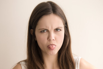 Fototapeta premium Headshot portrait of offended young woman. Angry teen girl with grumpy facial expression looking at camera with sticking out tongue. Cute female showing her contempt or disgust. Close up. Front view