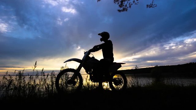 Silhouette of an extreme transport driver who sits on a motorcycle, a vehicle stands near a river on a sunset background