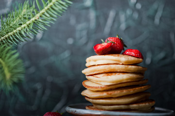 Beautiful homemade pancakes with strawberries and raspberry red jam on a black wall background. Pancakes with fresh juicy strawberries and a vase with a sprig of Christmas trees. Pancakes watered with