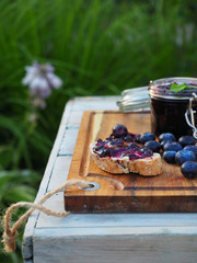 Homemade blueberry jam in glass jar with mint leaves. Piece of bread with butter and blueberry jam on cutting board. White wooden table and green grass on background.