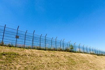 Fototapeta na wymiar A border made of a double wire fence with barbed wire on top of a bank under a blazing sun and a summer blue sky.