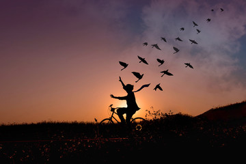 Freedom concept, Silhouette of happy person raised arms on bicycle in natural scene, Birds fly on beautiful sunrise or sunset sky