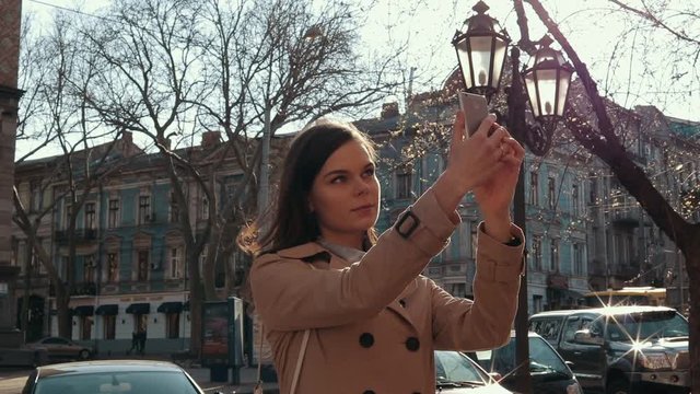 Brunette woman in a beige coat takes pictures of herself on a smart phone in the city in the spring, slow motion