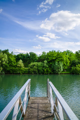 Naklejka na ściany i meble A wooden pontoon with metallic handrail over a river with trees and vegetation under a blue sky with white clouds.