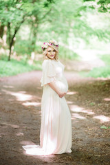 Fototapeta na wymiar Pregnant woman with flower wreath poses in white dress in the park
