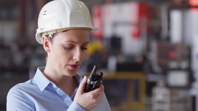 Slow motion shot of female engineer in hardhat talking on portable radio set and pointing somewhere while controlling manufacturing process at factory