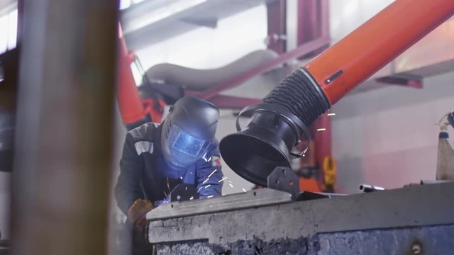 Tracking of factory worker in protective mask welding metal part of industrial machine on factory. Slow motion shot