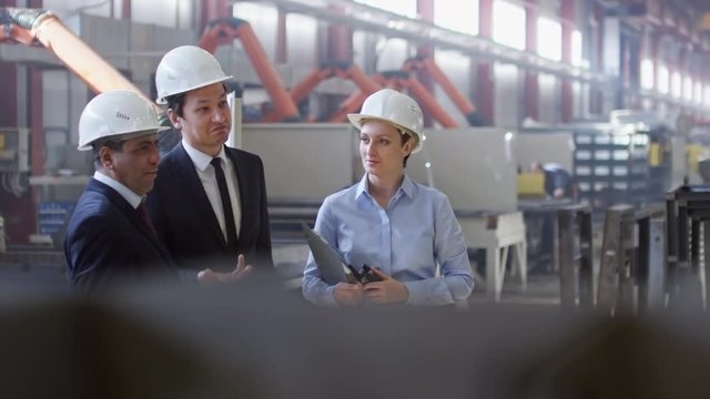 Slow motion shot of Asian and Latin-American engineers standing with female colleague in factory workshop,talking about something and then walking around.Woman holding documents and portable radio set