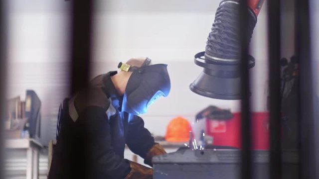 Side of industrial worker in protective uniform and mask welding something in factory workshop in slow motion