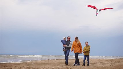 A young family walks along the beach next to the sea in cold weather, the son is holding a kite in his hands, the father carries a child, the mother is happy with a good weekend