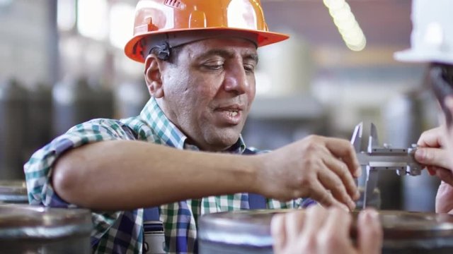 Tracking shot of experienced engineer using caliper for measuring metal detail and talking to colleague