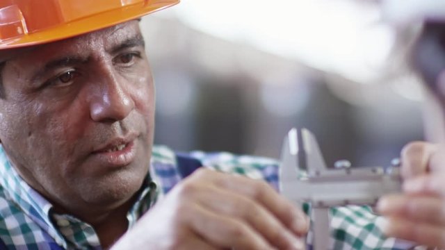 Closeup of Hispanic factory worker in hardhat measuring detail with vernier caliper and talking to somebody