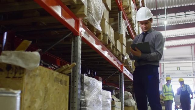 Male supervisor standing in hardhat in factory warehouse with clipboard and checking boxes on shelves