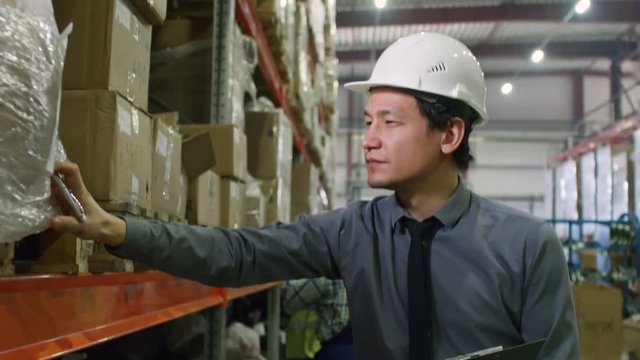 Tilt down of factory supervisor standing in hardhat in warehouse, checking boxes on shelves and writing on clipboard