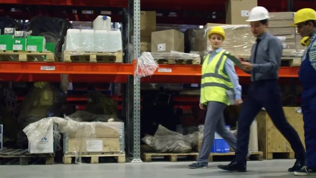 Side view of factory supervisor walking with two colleagues along shelves in warehouse in slow motion