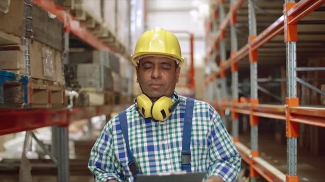 Latin American factory worker in hardhat walking in slow motion along shelves in warehouse with protective headphones over neck and writing on clipboard