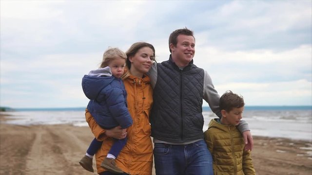 A happy and friendly family is walking with their young children on the beach near the sea on a day off, mom is carrying a baby, a middle-aged man is hugging his wife and son