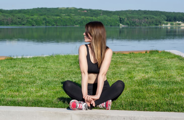 Fototapeta na wymiar Slim girl sitting on the grass on the shore of the lake. Fitness on the coast.