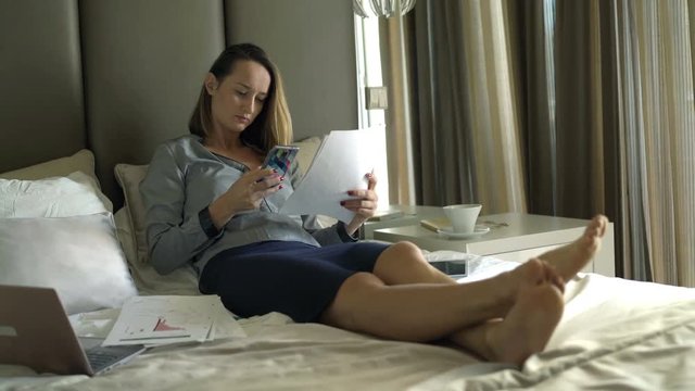 Businesswoman counting data from documents on smartphone lying on bed at home
