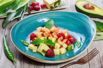 Fruit salad with shrimp, avocado, bulgarian pepper, kiwi, pineapple, raspberries in plate on wooden background close up. Mediterranean food. Top view