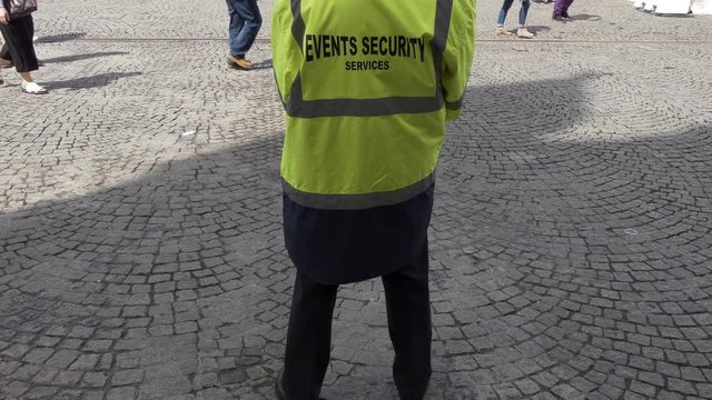 Rear view of security man wearing high visibility coat standing in pedestrian area outdoors. Unrecognizable people walking around, some woman comes to him to ask a question.