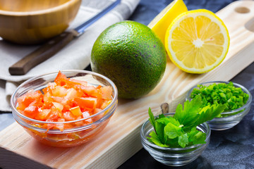 Fresh ingredients for guacamole on dark background.  Tomatoes, lemon, chives, parsley, avocado.