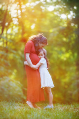 The girl hugs her pregnant mother at the edge of the forest. Woman and daughter are standing in the evening haze and the golden rays of the sunset