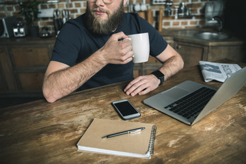 cropped view of bearded freelancer drinking coffee and working with laptop in home office