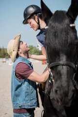 smiling woman in helmet sitting on horseback and looking on young man in cowboy hat