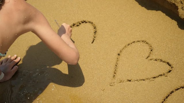 Girl on a beach draws a hearts on sand.

