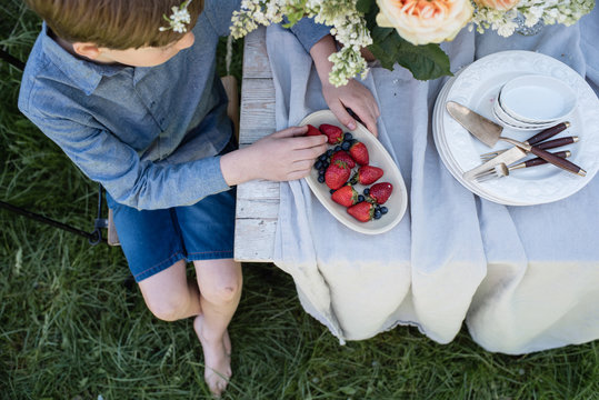 Junge sitzt barfuß  im Garten am Tisch und isst Erdbeeren